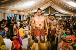 Young shirtless Samoan male dancers in traditional outfits standing amidst Indian guests in decorated canopy tent during wedding party